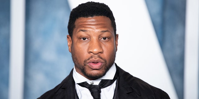 Jonathan Majors looks directly at the camera wearing a black suit, white shirt and slightly loose black tie at the Vanity Fair Oscar party