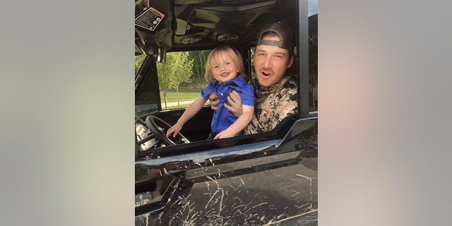 Morgan Wallen in a camo shirt and backwards hat holds his son in a blue shirt in his car