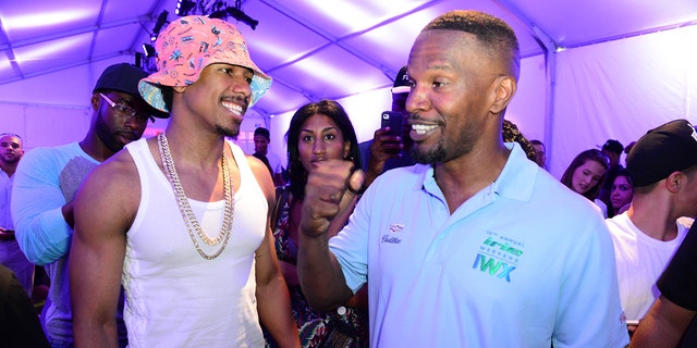 Nick Cannon in a pink hat, a white tank top and large chain smiles at Jamie Foxx who has his fist in the air wearing a blue collared shirt