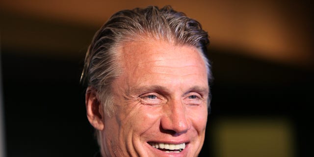 Dolph Lundgren smiles wearing black button down shirt