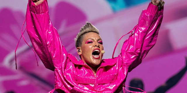 Pink throws her arms up in the air wearing a hot pink latex-like jacket while on stage