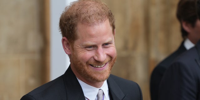 Prince Harry smiles in a black suit and white button down with medals adorned on his suit at the coronation of King Charles