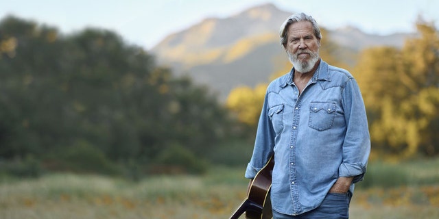 Jeff Bridges holds guitar while posing in the mountains 