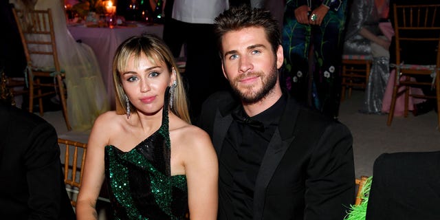 Miley Cyrus in a green and black gown sits at a table next to Liam Hemsworth in all black at The Met