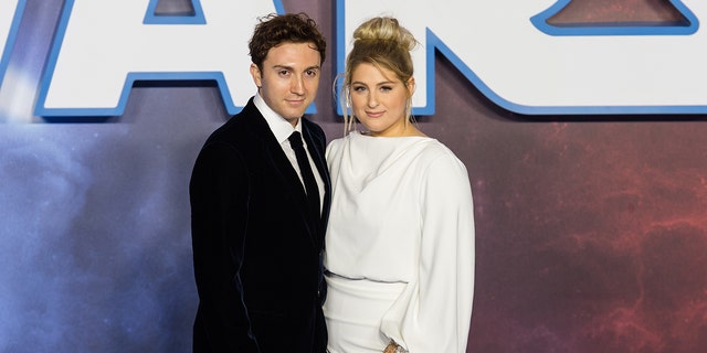 Daryl Sabara in a black suit and tie with a white shirt poses on the red carpet with wife Meghan Trainor with a glamorous up-do, long white gown, and hand bracelet