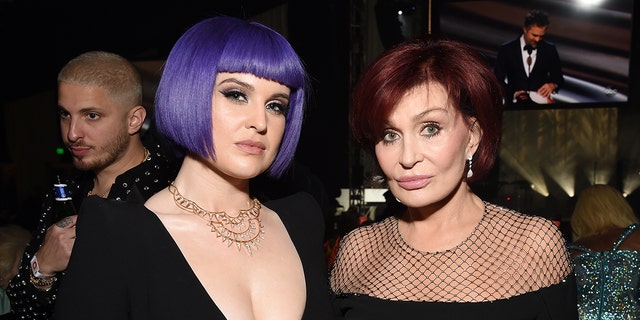 Kelly Osbourne with a short purple bob and deep V-neck black dress smiles for a photo with her mother Sharon in a black gown with a mesh top