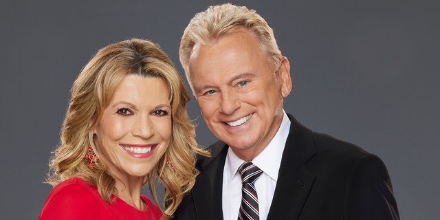 Vanna White in a red dress poses with Pat Sajak in a black suit and patterned tie for "Celebrity Wheel of Fortune" promo shots