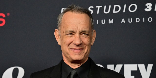 Tom Hanks smiles with his mouth closed wearing a black suit at the LA Convention Center in LA