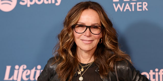 Jennifer Grey in a black leather jacket and black framed glasses smiles on the red carpet