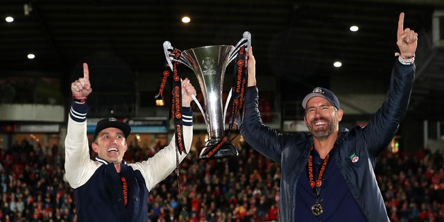 Rob McElhenney and Ryan Reynolds hold up trophy on field after Wrexham win.