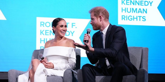 Meghan Markle wearing a white off the shoulder dress sitting next to Prince Harry in a suit and tie