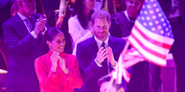 Meghan Markle wearing a red jumpsuit and Prince Harry wearing a blue suit in front of the American flag