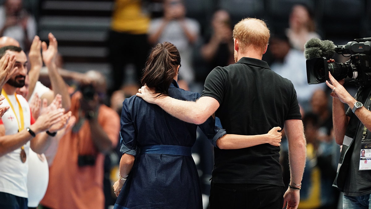 Meghan Markle wearing a blue dress and Prince Harry wearing an all black outfit with their backs turned