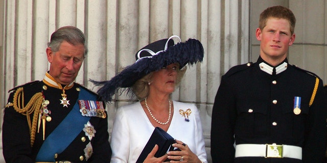 Prince Harry in a military suit looking annoyed as Camilla wears a white dress and an oversized navy hat standing next to prince charles in a military uniform