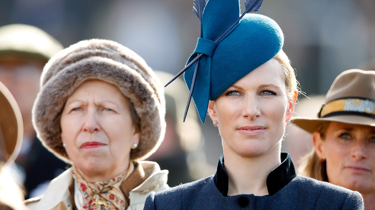 Zara Tindall wearing a blue coat dress with a matching hat as she stands next to Princess Anne weight a brown coat and a fur hat.