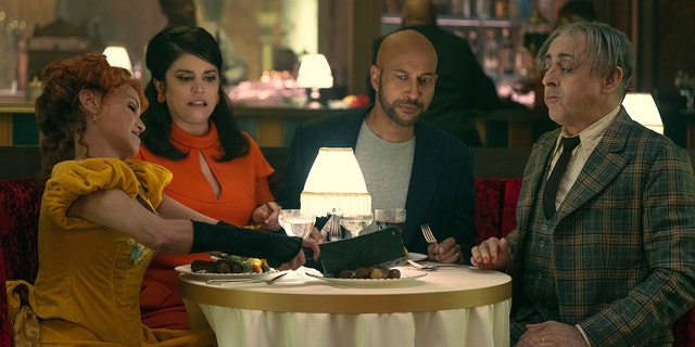 The cast of "Schmigadoon!" including Kristin Chenoweth in yellow, Cecily Strong in a bright orange, Keegan-Michael Key in a grey shirt and navy blazer and Alan Cumming in a green plaid suit set sit around a table in a photo from the second season