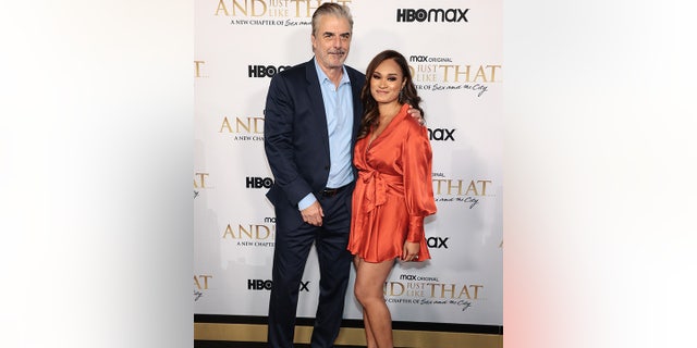 Chris Noth in a blue shirt and black suit smiles on the carpet with his wife Tara Wilson in a silky burnt orange short dress