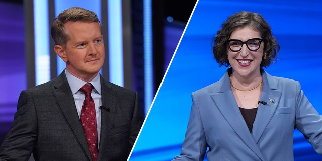 Ken Jennings in a dark suit and red tie looks to contestants while on Jeopardy! split Mayim Bialik in a blue jacket behind a podium on "Jeopardy!"