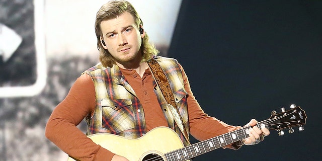 Morgan Wallen strums a guitar on stage while wearing plaid vest and jeans