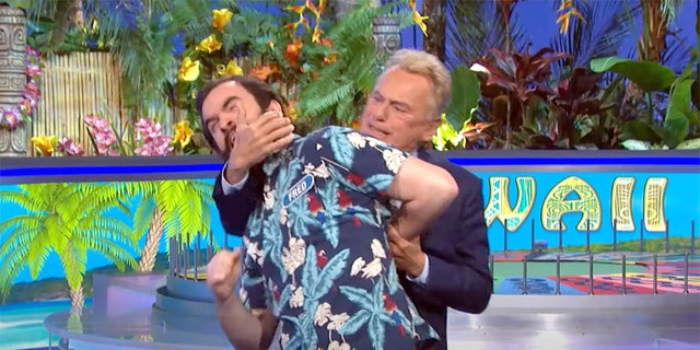 Contestant Fred in a patterned blue shirt has his arm locked behind him and neck locked by Pat Sajak as he jokingly is tackled